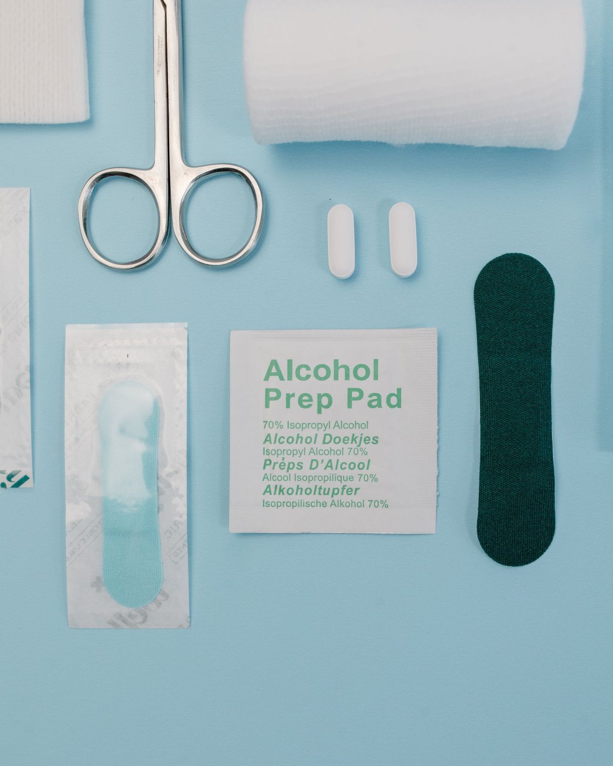 Vue de dessus sur fond bleu : compresse, ciseaux, bande, deux comprimés, pansement en sachet, lingette antiseptique et pansement vert